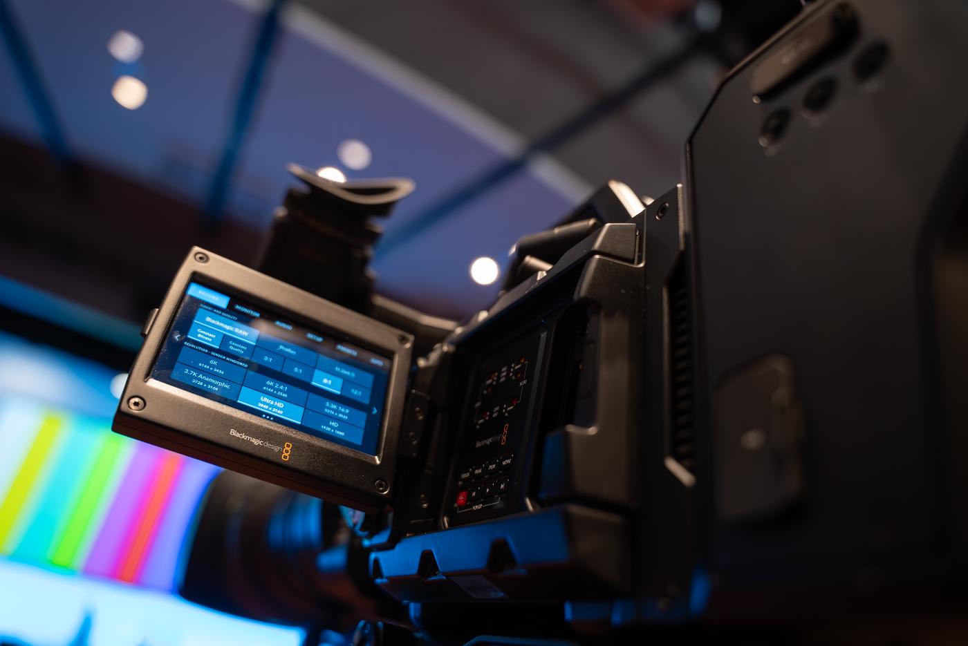 A close-up of a film camera, with the view finder flipped out. We can see that it's pointed at a stage, ready to record an event.